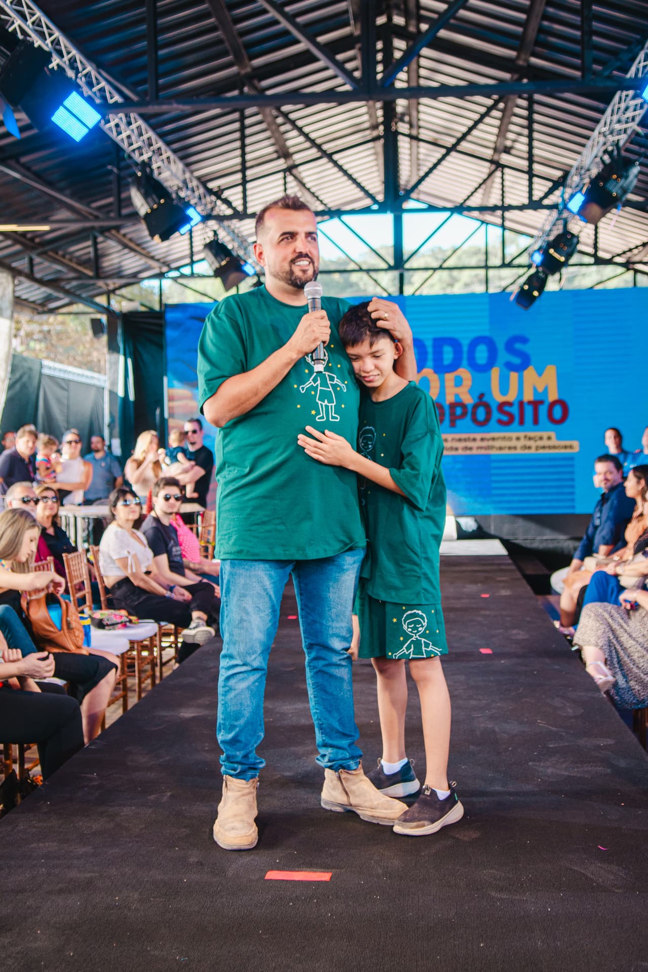 João abraçando o filho Enzo na passarela, com o painel do evento ao fundo